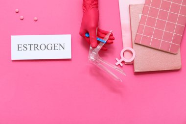 Doktorun elinde ESTROGEN sözcüğüyle jinekolojik spekulum, hormonal haplar ve pembe arka plan üzerine not defterleri var.