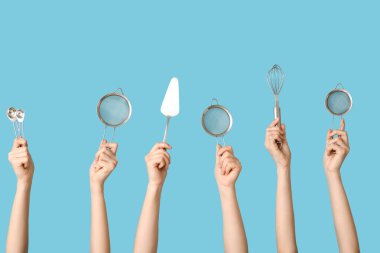 Kaşıklar, pasta spatulası ve mavi arka planda çırpılmış çırpılmış süzgeçleri tutan kadın eller.