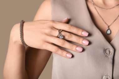Gri yüzüklü, bilezik takan ve kolyesi bej arka planda olan şık bir kadın.
