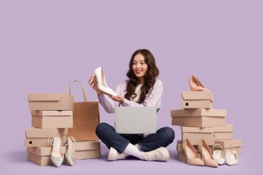 Laptop ve ayakkabı kutularıyla genç Asyalı kadın leylak arka planda alışveriş yapıyor.