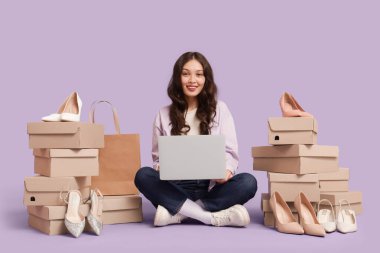 Laptop ve ayakkabı kutularıyla genç Asyalı kadın leylak arka planda alışveriş yapıyor.