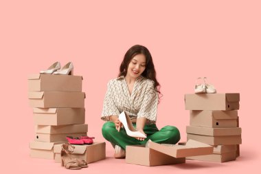 Pembe arka planda oturan yüksek topuklu ve ayakkabı kutularıyla genç Asyalı kadın.