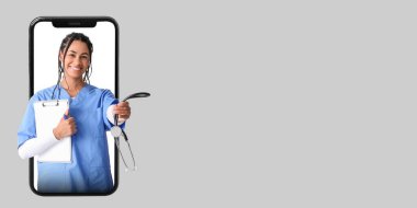 Steteskoplu genç Afro-Amerikalı bayan doktor gri arkaplandaki akıllı telefon ekranından mesaj için yer arıyor.