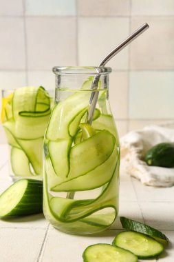 Beyaz fayans masasında salatalık dilimleri ile doldurulmuş şişe ve bardak su.