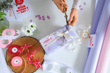 Hediye kutusu olan kadın eller, hafif arka planda paketleme malzemeleri ve çiçekler. Uluslararası Kadınlar Günü