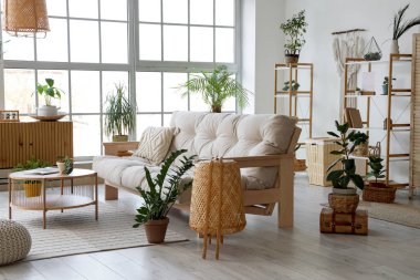 Rahat kanepesi, ahşap göğüs çekmecesi, masası, çiçekleri, raf ünitesi ve penceresi olan ışıklı şık bir oturma odası.