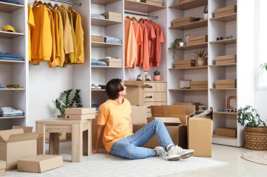 Giysi kutuları ve şık giysileri olan yakışıklı genç adam soyunma odasında oturuyor.