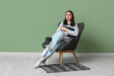 Yeşil duvarın yanında kitapla oturan genç bir kadın.