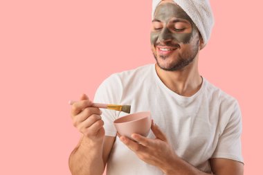 Yüzünde kozmetik kil maskesi olan yakışıklı genç adam pembe arka planda.