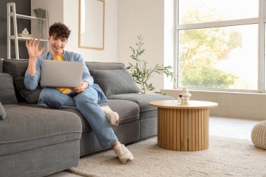 Genç adam evdeki kanepede dizüstü bilgisayarla görüntülü sohbet ediyor.