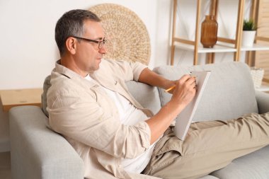 Yaşlı adam kanepede uzanıyor ve evdeki deftere bir şeyler yazıyor.