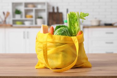 Mutfaktaki ahşap masada farklı taze ürünler bulunan sarı alışveriş çantası.