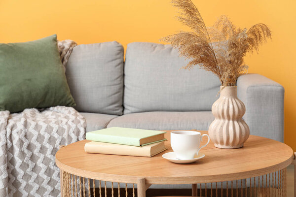 Cozy sofa, coffee table with pampas grass in vase, books and cup of tea near orange wall