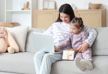 Genç anne ve küçük kızı hediye kutusuyla evdeki kanepede dizüstü bilgisayar kullanıyorlar.