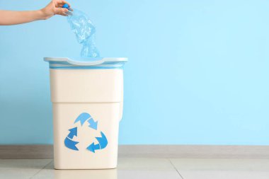 Kadın eli, mavi duvarın yanında geri dönüşüm logosu olan boş plastik şişeyi çöp kutusuna atıyor.