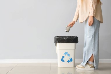 Gri duvarın yanında geri dönüşüm logosu olan çöp kutusuna konserve atan kadın.