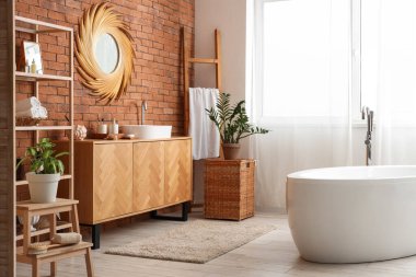 Tuğla duvarda asılı yuvarlak ayna, beyaz küvet, banyonun içinde cilt bakım ürünleri olan ahşap raf ünitesi.