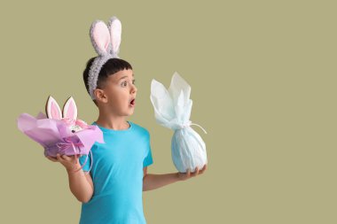 Şok olmuş küçük çocuk yeşil arka planda paskalya pastası ve hediye yumurtası ile tavşan kulağı takıyor.