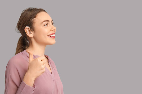 Красивая молодая женщина со слуховым аппаратом показывает большой палец вверх жест на сером фоне