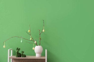 Ağaç dalları olan vazolar, yeşil duvarın yanındaki raflarda Paskalya yumurtaları ve tavşanlar.