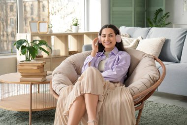 Kulaklıklı genç ve güzel bir kadın. Sesli kitap dinliyor ve oturma odasında koltukta oturuyor.