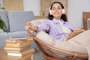 Kulaklıklı, kitapla sesli kitap dinleyen ve oturma odasındaki koltukta oturan güzel genç bir kadın.