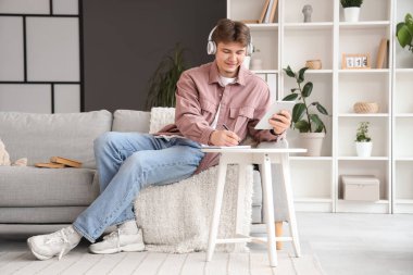 Kulaklıklı, tablet bilgisayarlı bir erkek öğrenci evde çalışırken not alıyor.