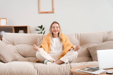 Genç bir kadın şık bir oturma odasında kanepede meditasyon yapıyor.