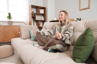 Genç bir kadın şık bir oturma odasında kanepede meditasyon yapıyor.