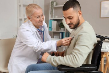 Steteskoplu kadın doktor hastanede tekerlekli sandalyedeki genç adamı dinliyor.