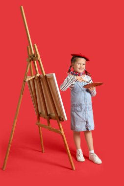 Kırmızı arka planda boya paleti ve sehpası olan küçük bir sanatçı.