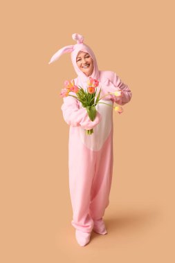 Paskalya tavşanı kostümlü, bej arka planda laleleri olan komik, son sınıf öğrencisi kadın.