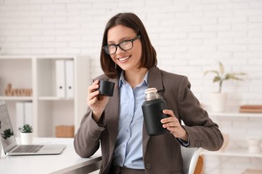 Işıl ışıl ofisindeki çalışma yerinde termosu olan genç iş kadını