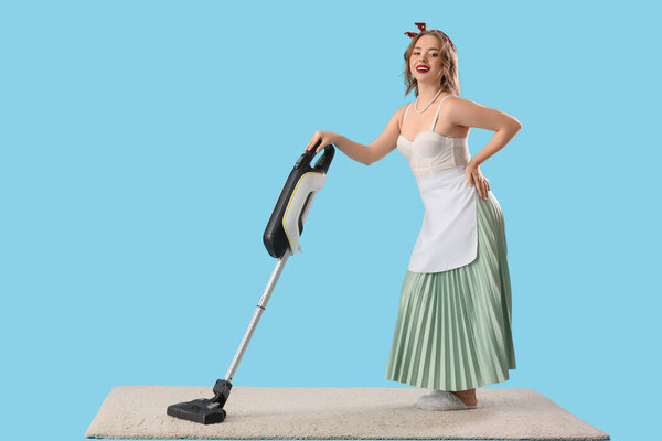 Young pin-up housewife vacuum cleaning carpet on blue background
