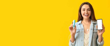 Sarı arkaplanda cep telefonu ve elektrik bankasını tutan mutlu genç bir kadın.