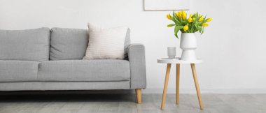 Oturma odasındaki gri kanepenin yanındaki masada sarı lale vazosu.