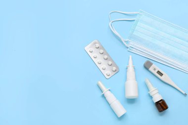 Mavi arka planda tıbbi maskeli, haplı ve termometresi olan burun damlaları.