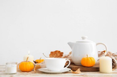 Çay demliği, bir fincan sıcak içecek, balkabağı, yanan mumlar ve sonbahar yaprakları beyaz duvarın yanında.