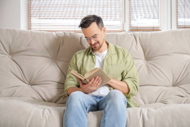 Gözlüklü yakışıklı genç adam evdeki kanepede kitap okuyor.