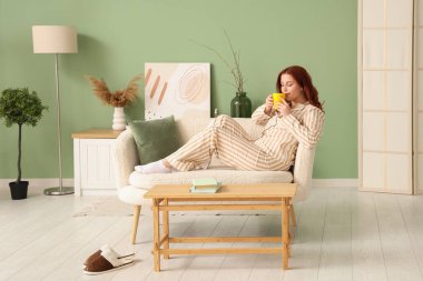 Pijamalı genç bir kadın evdeki kanepede çay içiyor.