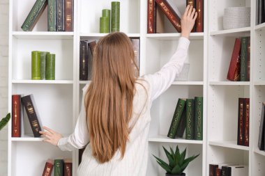 Çalışan genç bir kadın şık bir ofiste kitaplıktan kitap alıyor.