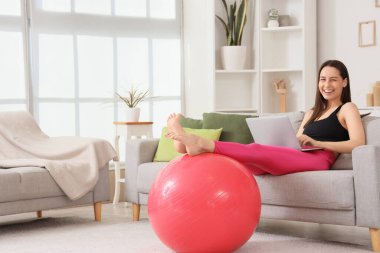 Evdeki spor salonunda dizüstü bilgisayar kullanan genç bir kadın.