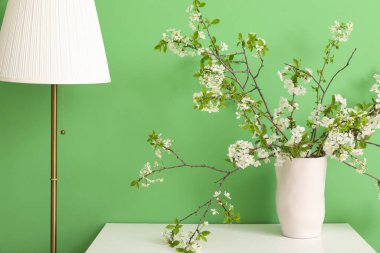 Yeşil odada, lambanın yanında, beyaz masanın üzerinde çiçek açan dalları olan vazo.