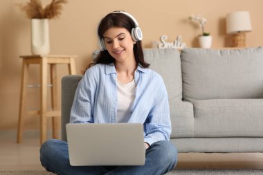 Genç bir kadın oturma odasında dizüstü bilgisayar ve kulaklıkla yerde çalışıyor.