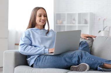 Genç bir kadın gri koltukta oturuyor ve şık bir oturma odasında dizüstü bilgisayar kullanıyor.