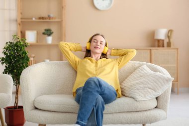 Kulaklıklı, müzik dinleyen ve şık oturma odasındaki kanepede dinlenen genç ve güzel bir kadın.