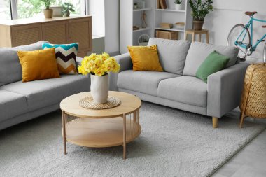 Aydınlık oturma odasının içinde gri kanepeler ve ahşap sehpanın üzerinde sarı narsisli çiçekler.