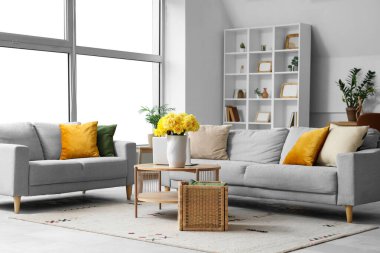 Oturma odasının içinde gri kanepeler, dizüstü bilgisayarlar ve ahşap sehpada sarı narsisli çiçekler.