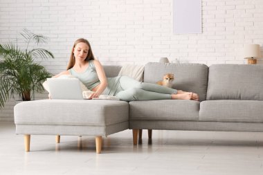 Evdeki kanepede dizüstü bilgisayarı ve sevimli kedisi olan genç bir kadın yatıyor.