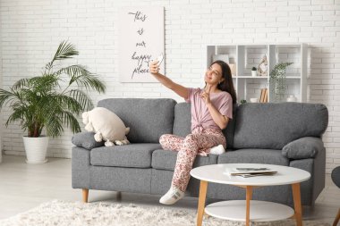 Oturma odasındaki koltukta cep telefonuyla selfie çeken genç bir kadın.